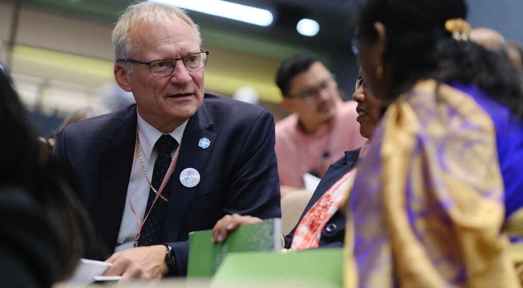 05 LWF generalforsamling Krakow Billedet viser en gruppe mennesker til konference i Det Lutherske Verdensforbund. Præsident Henrik Stubkjær bærer en mørk jakke med en hvid skjorte og en rød slips, og har badges fastgjort på venstre side af brystet. En anden person er iført traditionelt tøj med livlige farver. Baggrunden er sløret, men indikerer et indendørs miljø med kunstig belysning.
