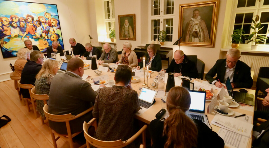 Toppix Stiftsråd Billedet viser stiftsrådet, der sidder omkring et ovalt træbord. De er engagerede i, hvad der ligner et møde eller en diskussion, hvor nogle kigger på dokumenter, og andre bruger bærbare computere. Forskellige genstande er spredt på bordet, herunder papirer, penne, briller og elektroniske enheder, hvilket indikerer en aktiv arbejdssession. Rummet er godt oplyst og har to store kunstværker på væggene; det ene er et farverigt maleri af flere figurer, mens de to andre er indrammede portrætter. Store vinduer lader naturligt lys strømme ind i rummet og forbedrer synligheden.