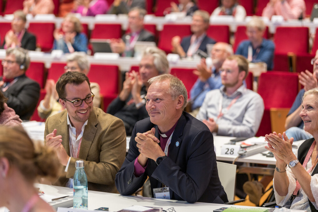07 LWF generalforsamling Krakow Billedet viser en gruppe mennesker, der sidder i et konferencelokale i Krakow i Polen med røde stole. Den kommende præsident Henrik Stubkjær sidder i midten af billedet. Nogle personer klapper, mens andre har hænderne sammen, muligvis i forventning eller applaus. Der er en flaske vand og papirer på bordene foran nogle af deltagerne. Rummet er godt oplyst og virker rummeligt.