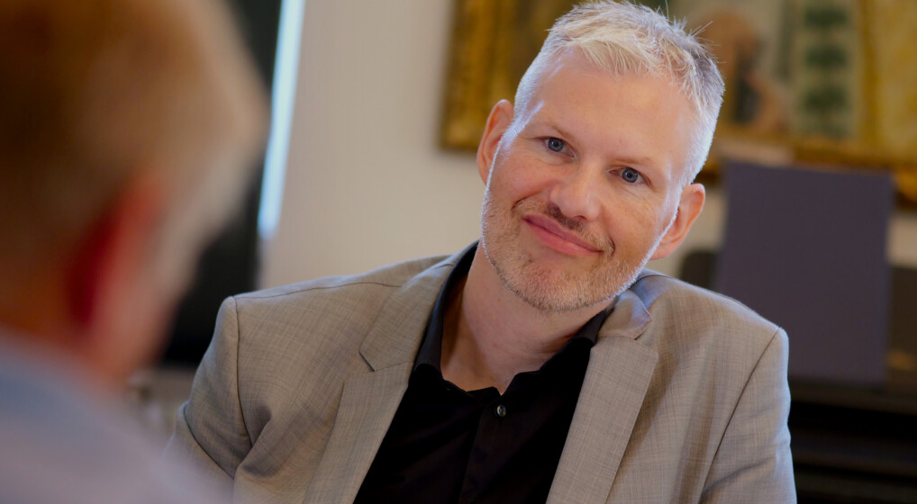 Stiftamtmand Jacob Hess i samtale med biskop Henrik Stubkjær. Jacob Hess vender mod kameraet, Han bærer en lys grå blazer og en sort skjorte. Henrik Stubkjær vises bagfra, og kun en del hans hoved og øre er synlige. I baggrunden er der kunstværker på væggen,